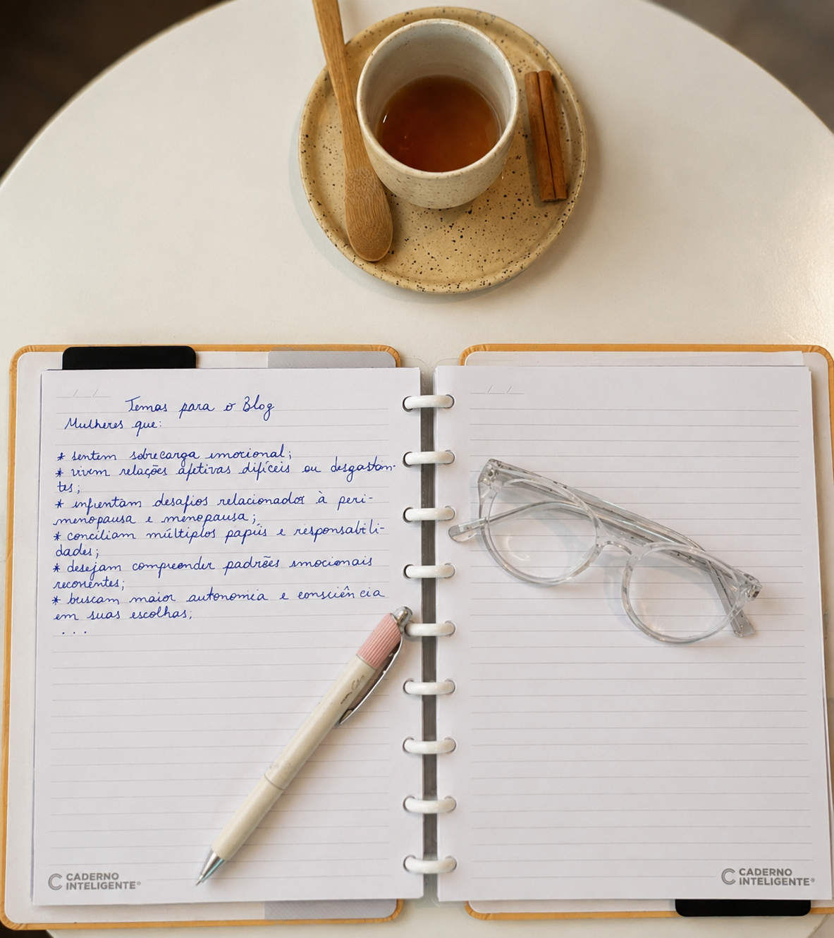 Caderno aberto com anotações, óculos e xícara de chá sobre mesa de café